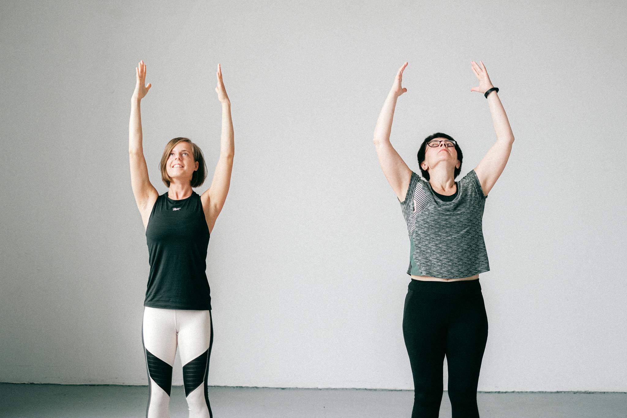 Two women engaged in a yoga session indoors, standing with arms raised, promoting fitness and wellness.